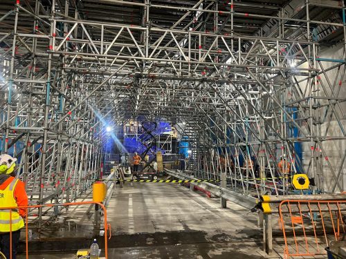 Layher Allround scaffold birdcage forming an access tunnel on a construction site, with a worker in hi-vis