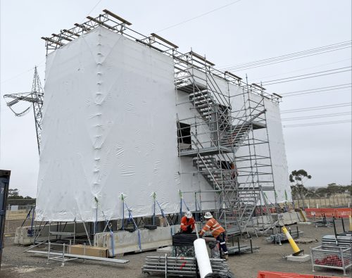 Crew working beside a shrink-wrapped scaffold enclosure with stair access, with stacked scaffold components nearby
